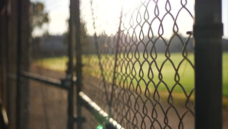 Close-up-of-a-chain-link-fence-gate-with-locks-on-it-at-sunrise-outside-of-a-grass-baseball-field-in-a-public-park