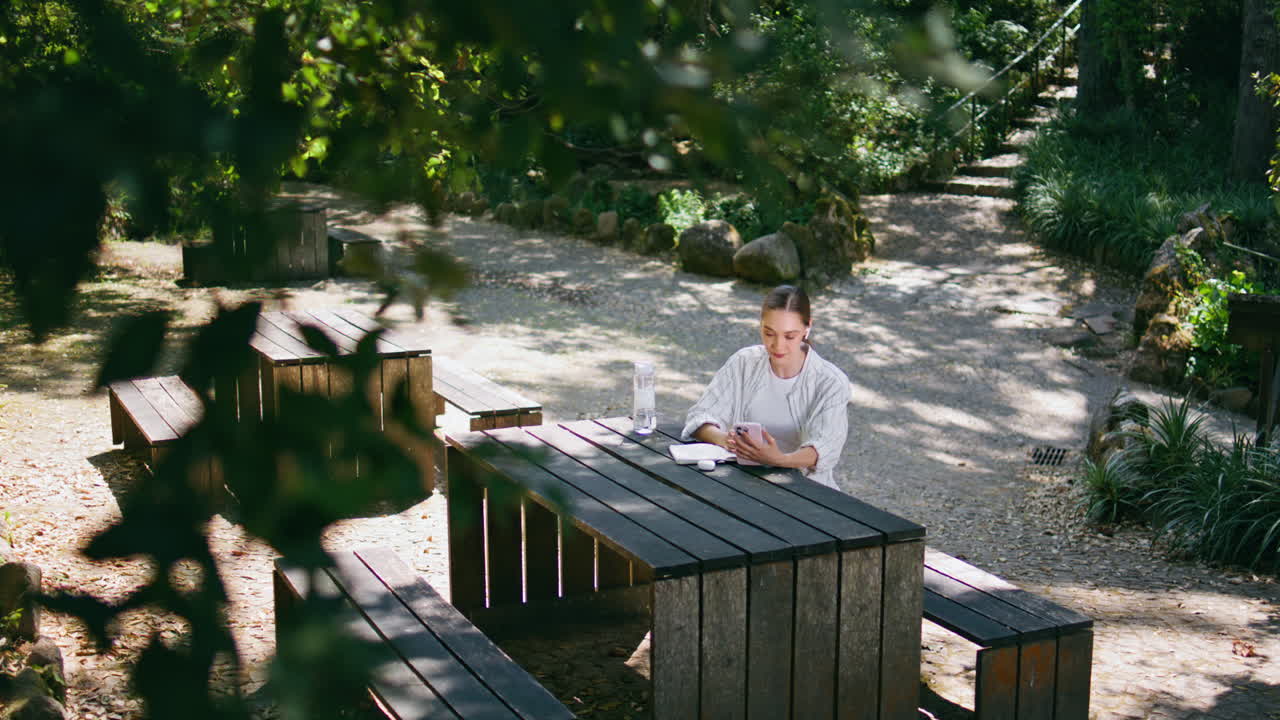 무선 헤드폰으로 음악을 듣는 진정된 여성이 테이블 숲에 앉아 있습니다.