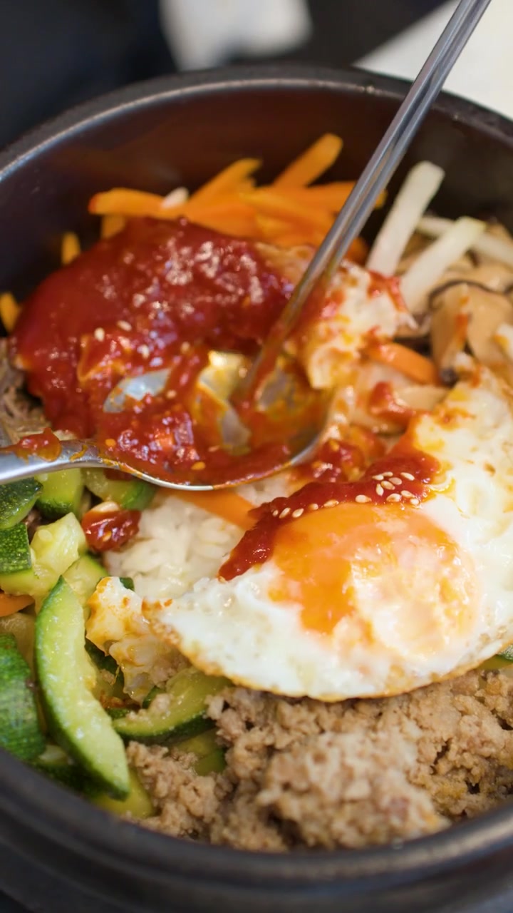 Bidikan jarak dekat sumpit dan sendok mengaduk bahan bibimbap dalam mangkuk batu panas.