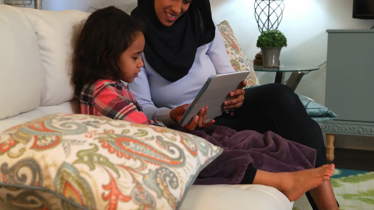 Young mother using a digital tablet with her daughter on the sofa at home 4k