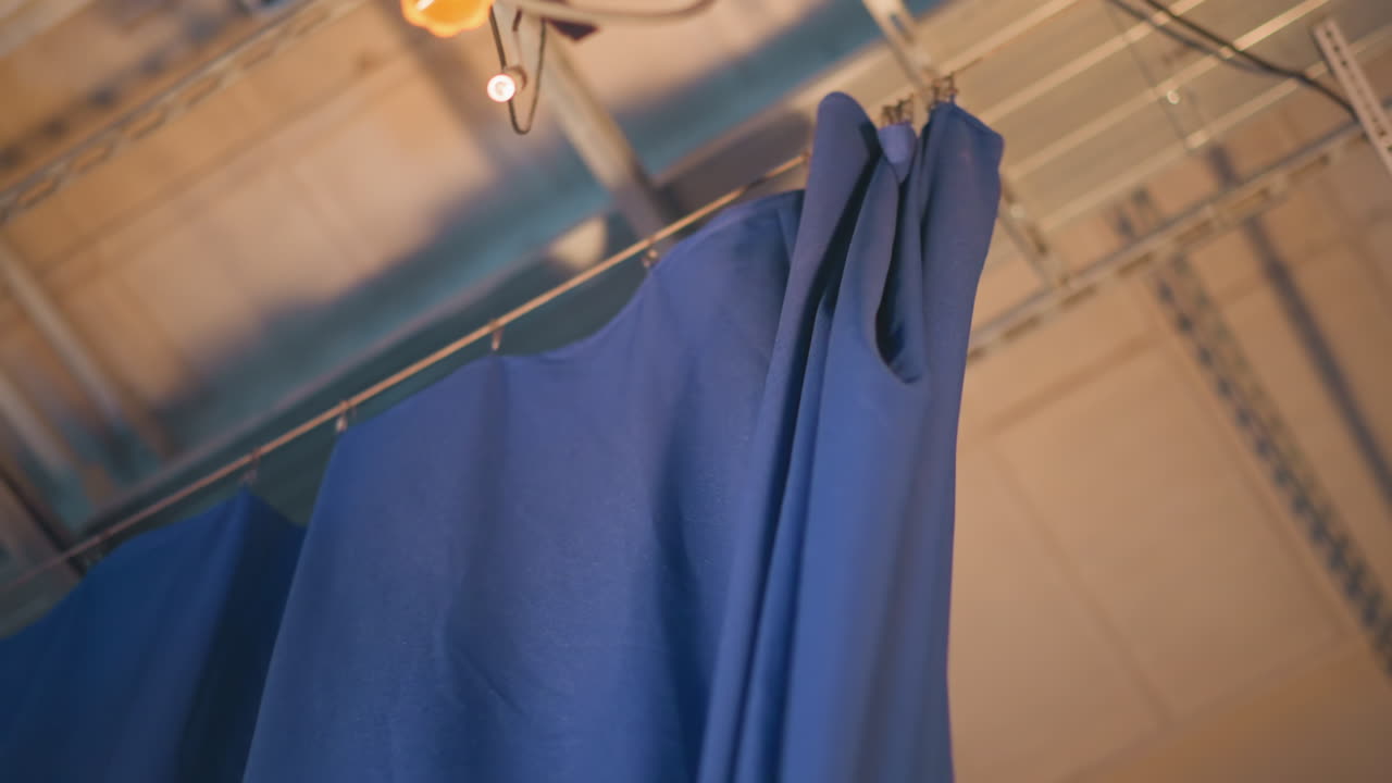 Male operator pulls blue curtain inside television studio set during production setup, floor lights flare below, overhead rigging visible, revealing striped backdrop ready for live news recording