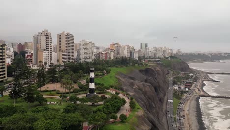 Aerial-orbital-shot-showing-a-city-coast-lighthouse-in-the-middle-of-a-park-with-people-while-cars-are-driving-below-on-a-highway-next-to-the-ocean-and-paragliders-flying-above