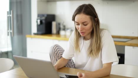 Blonde-woman-is-making-online-payment-holding-bank-card-using-modern-laptop-at-home