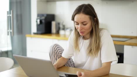 blonde woman is making online payment holding bank card using modern laptop at home