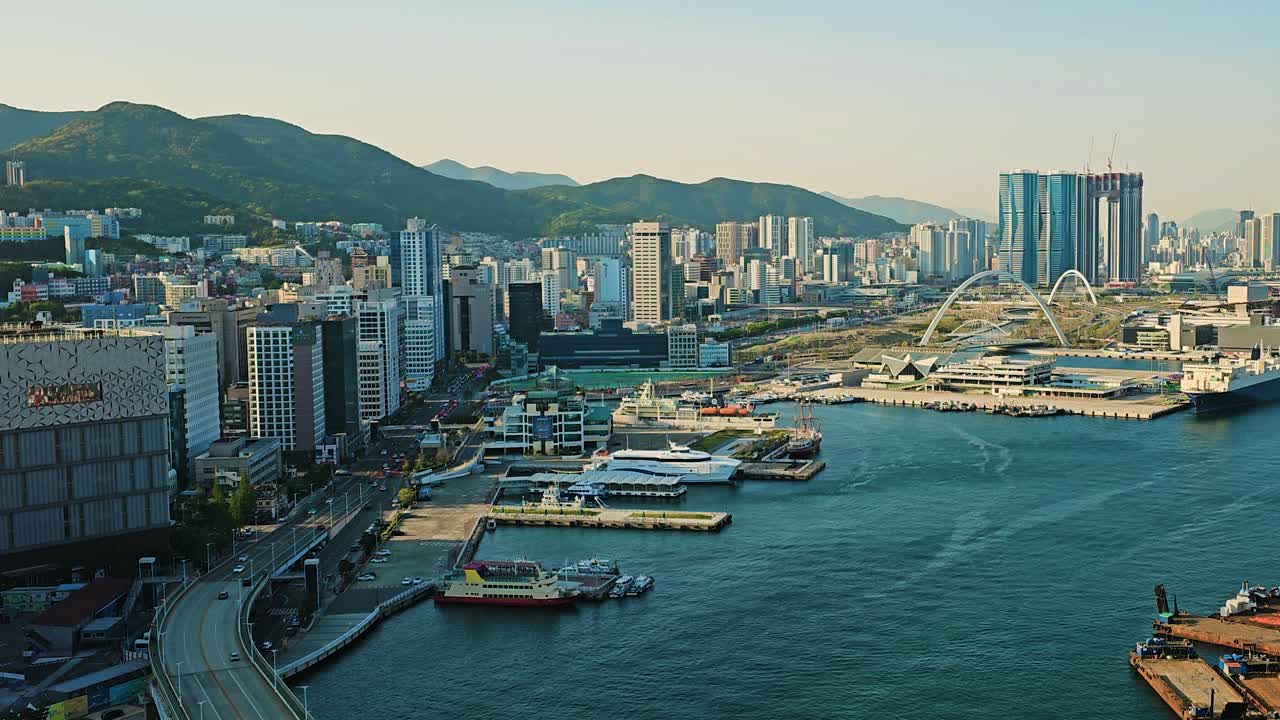 Wide aerial view of Busan's coastal cityscape with modern high-rises, harbor docks, ships, and surrounding green mountains during golden hour