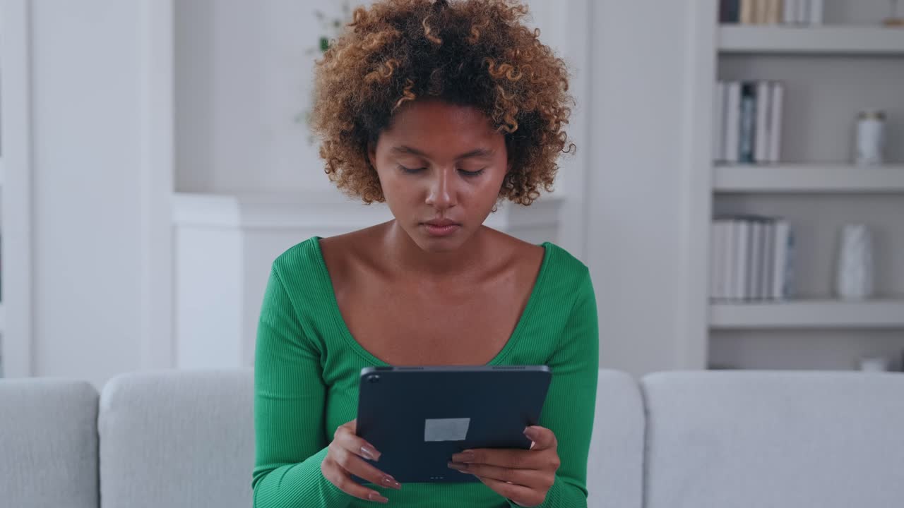 Young alarmed african american woman watches video on tablet sits on sofa