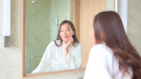 asian woman in hotel bathrobe checking and touching her pretty face near the mirror in the bathroom slow-motion