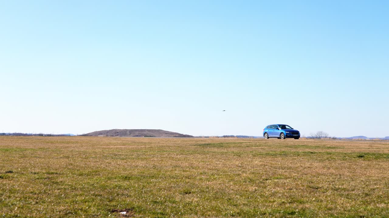 Drone Capturing Aerial Footage of a Car Driving Across an Expansive Field Under a Clear Blue Sky