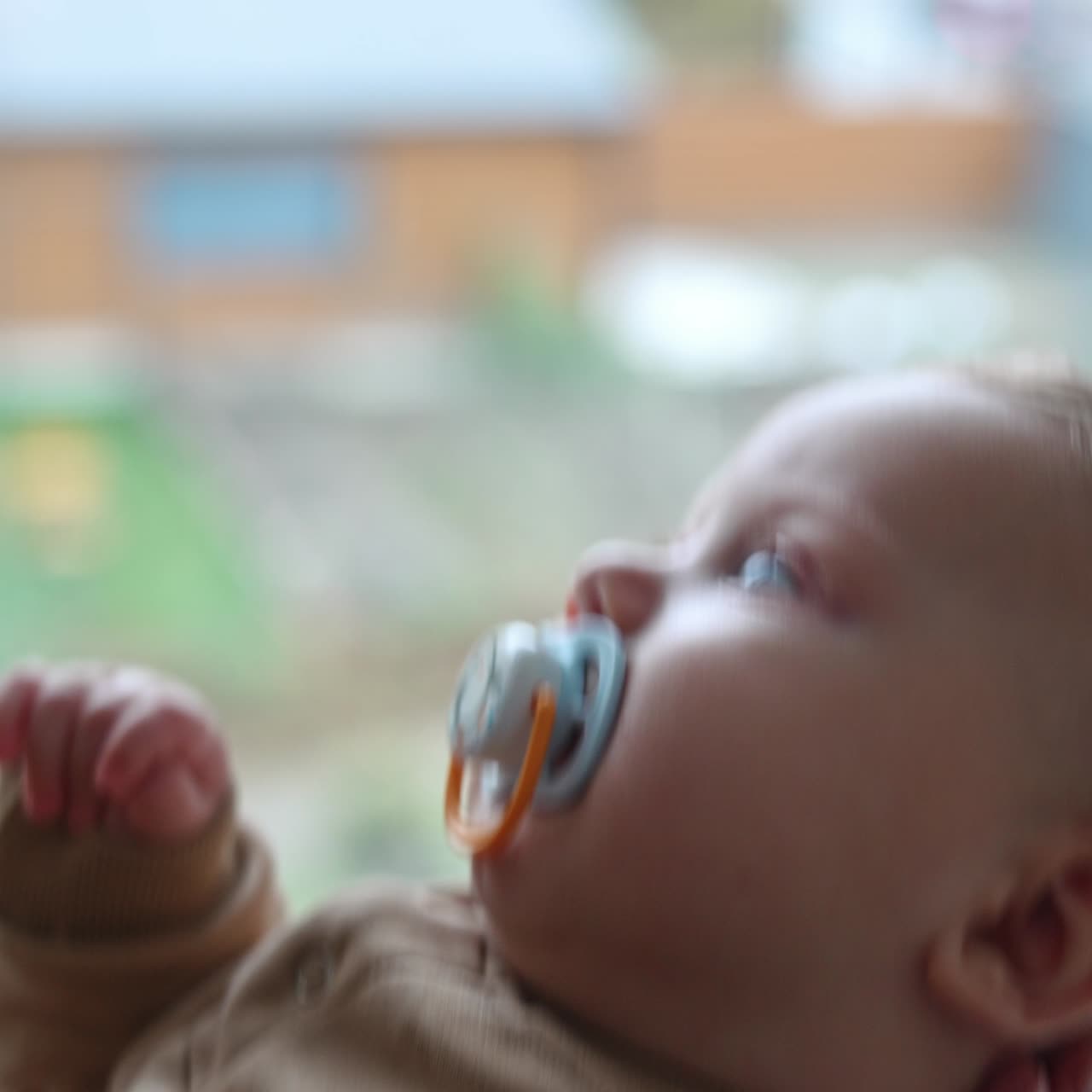 Unrecognized woman waving her cute child near the window. Calm baby boy with pacifier in mouth looks up