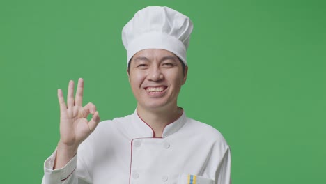 primer plano de un chef asiático sonriendo y mostrando un gesto de acuerdo a la cámara mientras está de pie en el estudio de fondo de la pantalla verde