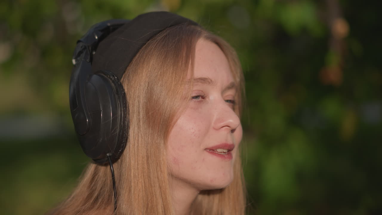 Parco illuminato dal sole, donna bianca con cuffie beve un frullato da uno shaker trasparente, chiude gli occhi, sorride al ritmo di musica lofi, atmosfera estiva rilassata, berretto e cuffie over-size, sfondo bokeh verde, ritratto spontaneo