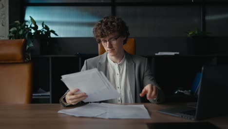 desde el lado un joven confiado con cabello rizado y gafas en una chaqueta gris examina documentos y papeles mientras está sentado en una mesa en una oficina moderna