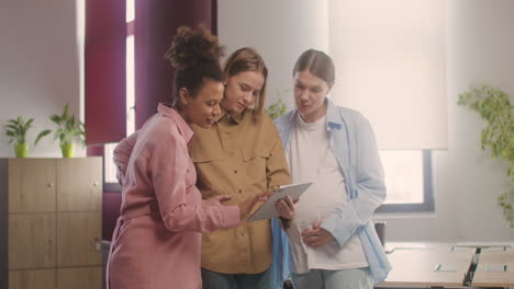 Three-Pregnant-Woman-Looking-At-Tablet-And-Talking-In-The-Office