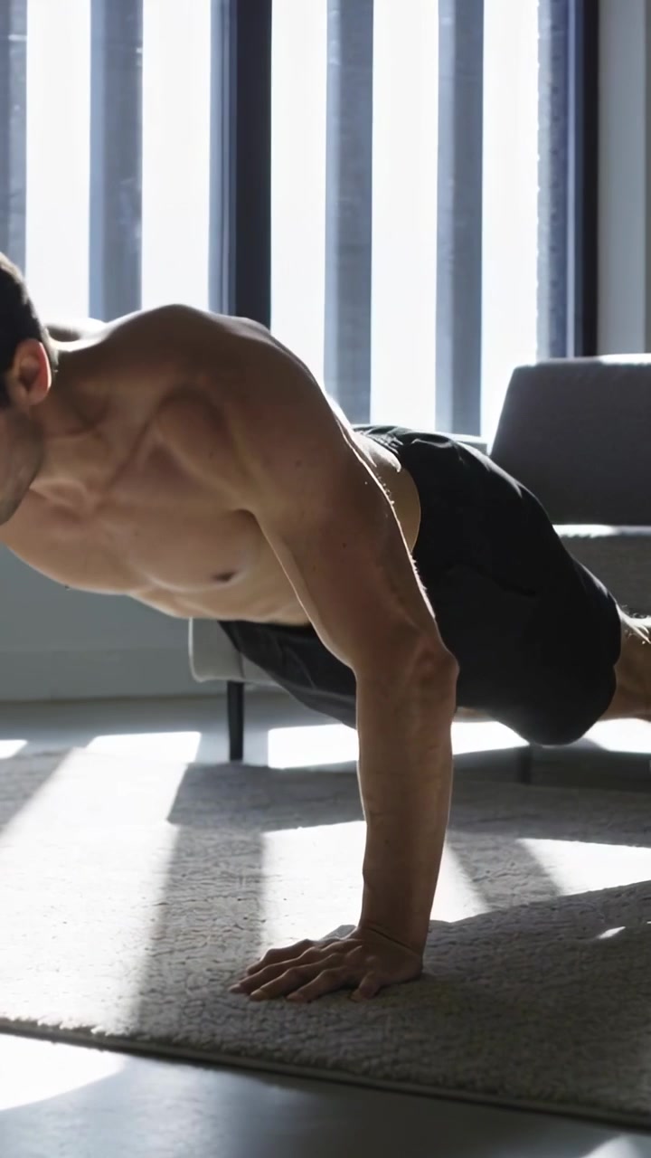 Video verticale: Uomo a torso nudo fa flessioni su un tappeto a casa, illuminato dal sole che filtra dalle persiane, per allenarsi.