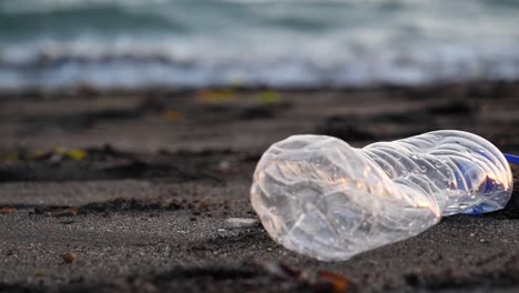 Zeitlupenaufnahme-Einer-Weggeworfenen-Plastikflasche-An-Einem-Sandstrand,-Während-Die-Wellen-Im-Hintergrund-Anspülen