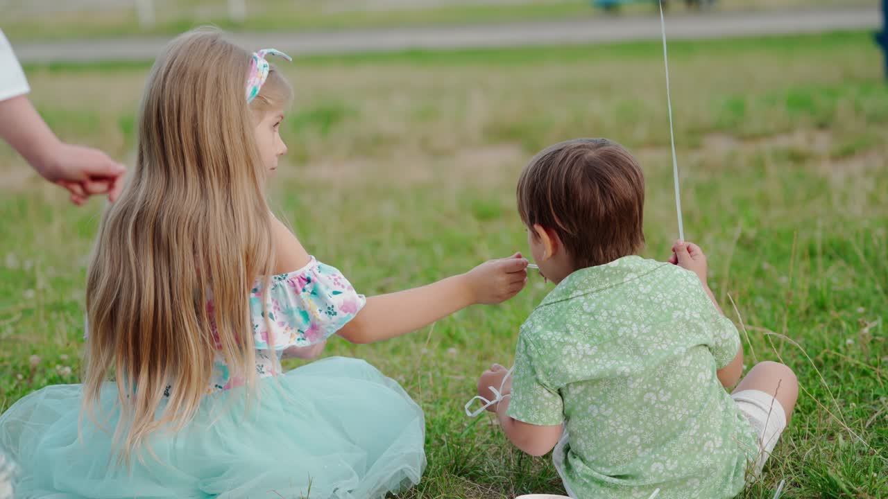 Backside view of a baby boy and a girl sitting on grass. Little sister feeding her smaller brother with a spoon outdoors.