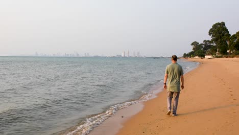 male tourist walking away along the beach of bangsaray in pattaya, thailand with city view in the distance