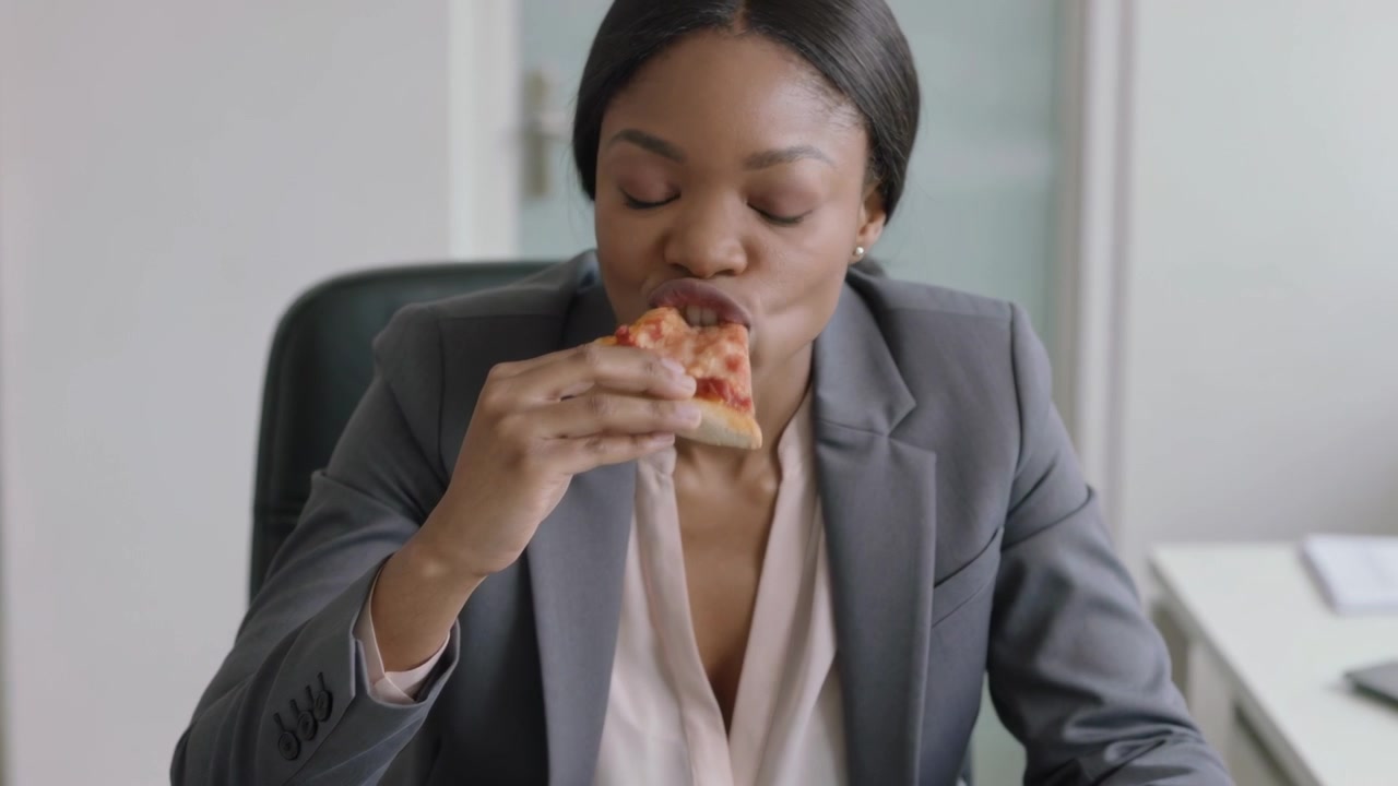 Holding woman in grey blazer bringing slice to mouth, biting during lunch at office with laptop