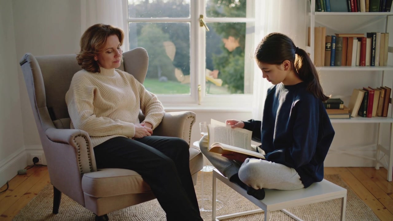 Una ragazza adolescente apre un grande libro, girando le pagine e condividendo una storia a casa, con la madre che guarda da una poltrona.