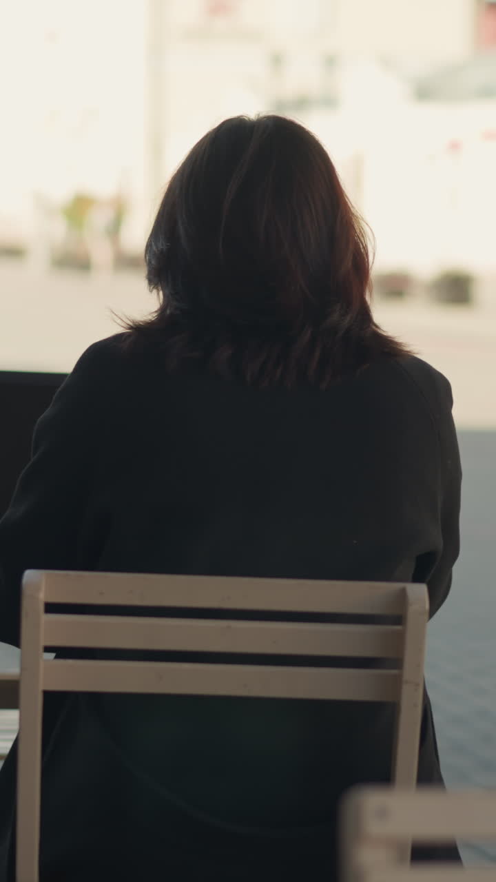 야외에서 노트북으로 일하는 여성의 뒷면, 머리카락이 얼굴을 고, 근처의 장식적인 검은 기둥, 현대적인 건물을 특징으로하는 흐릿한 도시 배경, 그리고 멀리 걸어가는 사람들