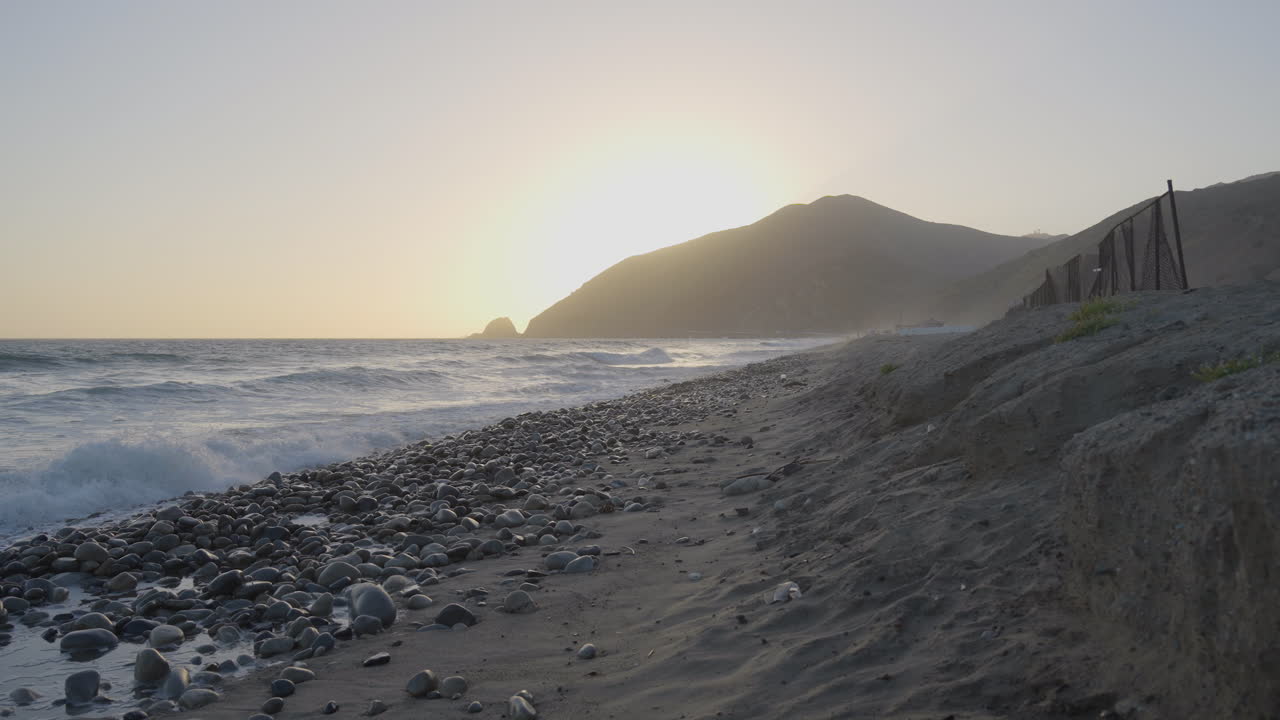 descendiendo a una toma panorámica que sigue la playa hasta el océano, donde la costa rocosa se convierte en el centro de atención con las olas rompiendo en la costa al fondo al atardecer en el sur de california