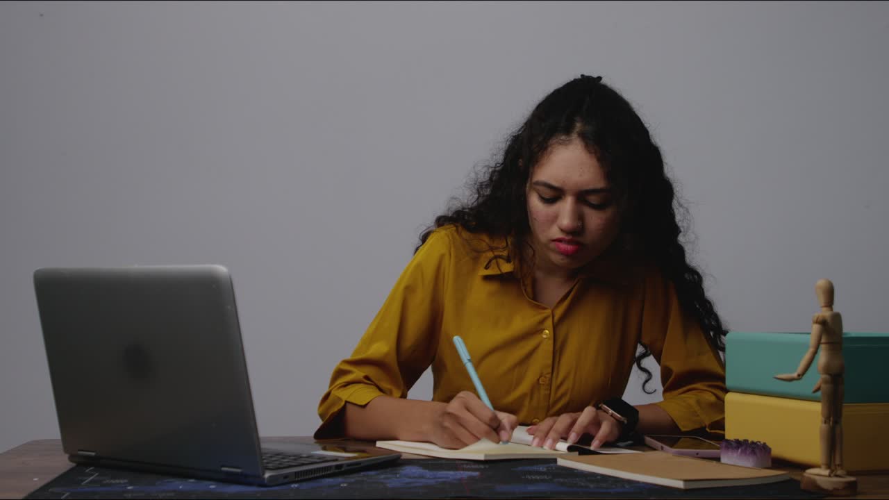 An engaged South Asian model writing notes in a notebook at her desk in a modern office setting, focused and immersed in thought