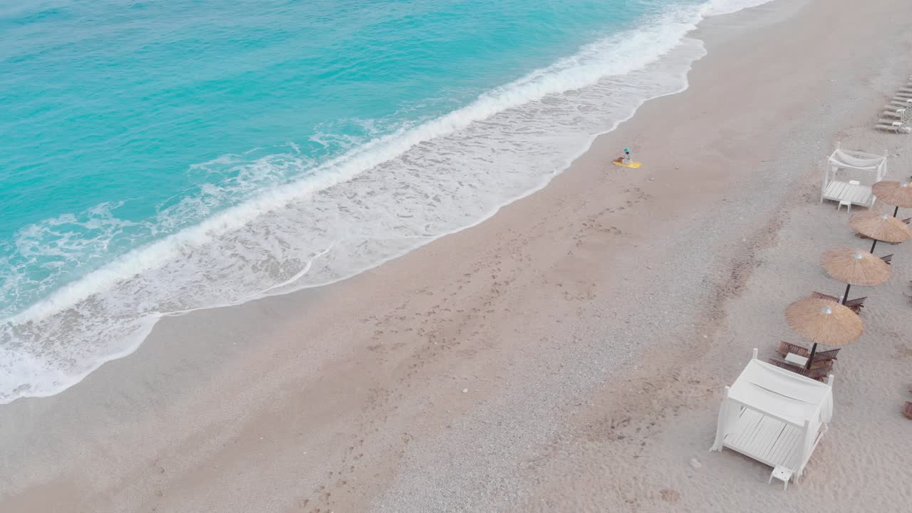 Kumlu okyanus plajında oturmuş ve deniz dalgalarına bakmakta olan bir kadının havadan drone görüntüsü.