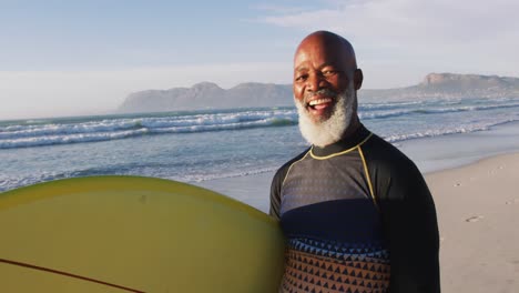 smiling senior african american man walking with a surfboard at the beach
