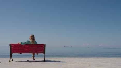 woman sits relaxed on a bright red bench, facing the calm blue adriatic sea in durrës, albania. large cargo ship is visible in the distance under a clear sky