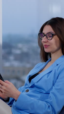 calm relaxed beautiful lady wearing blue jacket sits in armchair. woman looks at her phone, then takes off her glasses and smiles to camera. close up. vertical video