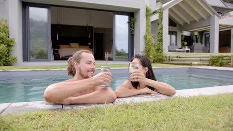 Una-Pareja-Diversa-Disfruta-De-Bebidas-Junto-A-La-Piscina,-Sonriendo-Y-Relajada