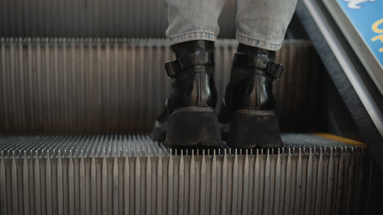 leg back view of female artist standing on moving walkway in mall wearing jeans and black boots captured from close angle showing grooved metal tread under bright lights reflective surface