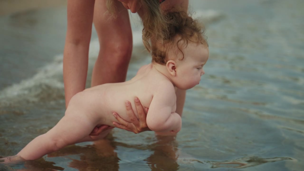 Imagens em close-up de um bebê adorável de cabelo cacheado explorando as águas rasas do mar, com o apoio da mãe, na praia.