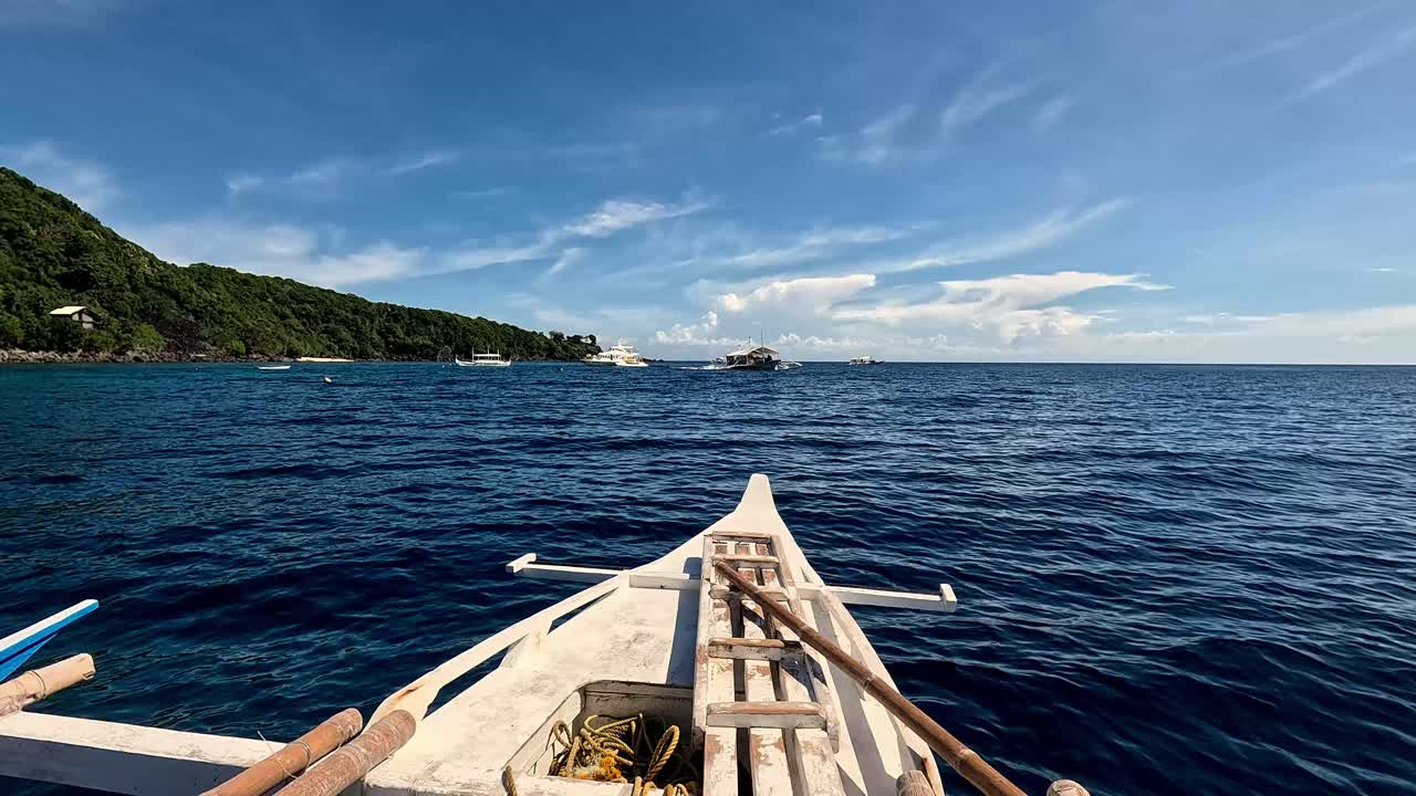 フィリピン、ダウインでの夏の旅行休暇。波打つ青い海をアウトリガーボートでセーリングするPOV。遠くの島と地平線に停泊中のボートが見える。