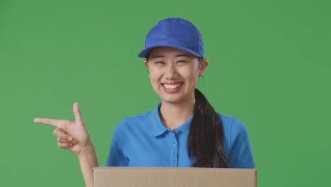 푸른 유니폼을 입은 아시아 여성 택배기사의 근접 사진. 미소 짓고 으로 가리키며 스튜디오에서 녹색 스크린 배경에 상자를 배달하고 있습니다.