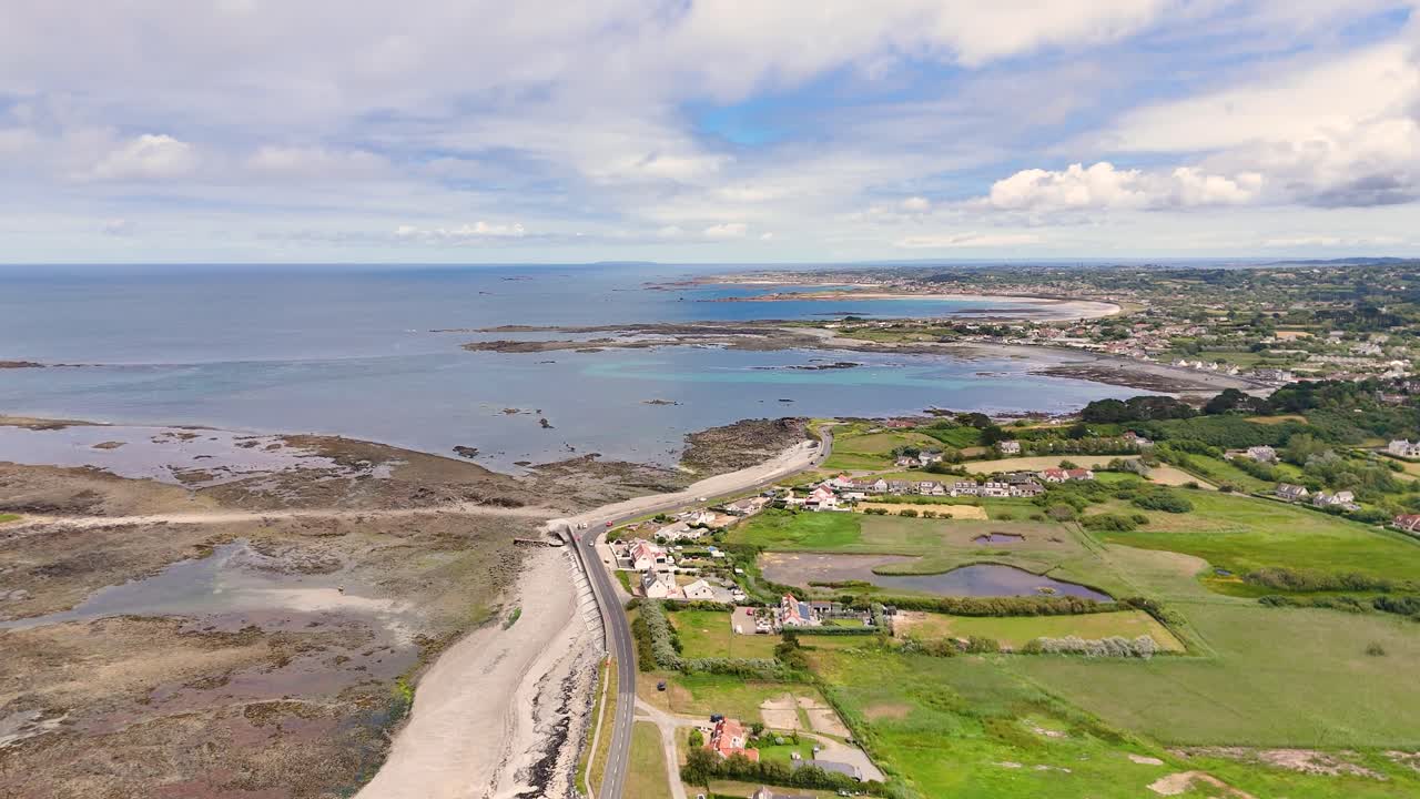 Guernsey. High flight along coast road on west coast of the Island over rocky foreshore with clear calm water towards headlands and distant golden beaches on sunny day