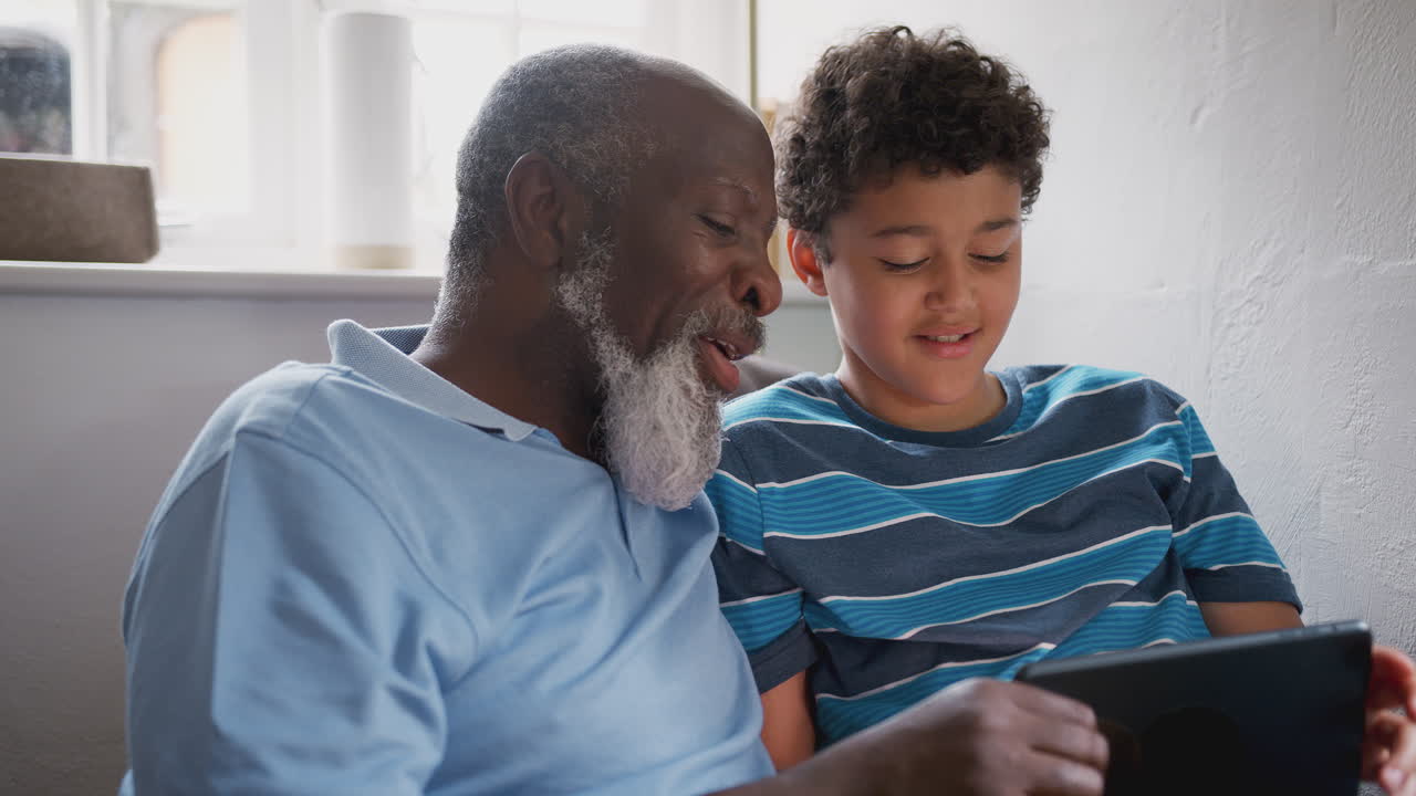 Grandfather Sitting In Chair As Grandson Explains How To Use Digital Tablet