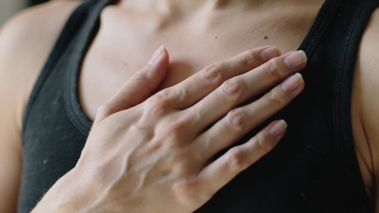 Donna in canotta nera che mette la mano destra sullo sterno, controllando il battito cardiaco sotto la luce della finestra.