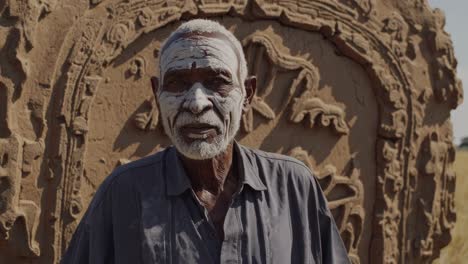 dogon elder wearing ceremonial white face paint standing near ancestral stone carved with symbolic cosmological markings in rural tireli village, mali's traditional cultural landscape