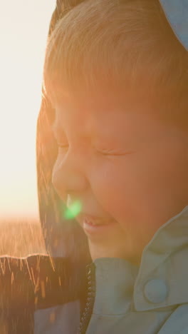 kid squints eyes under rain sprays outdoors. happy little boy in hooded raincoat stands under sunshower in sunset meadow closeup. beauty of rainy weather