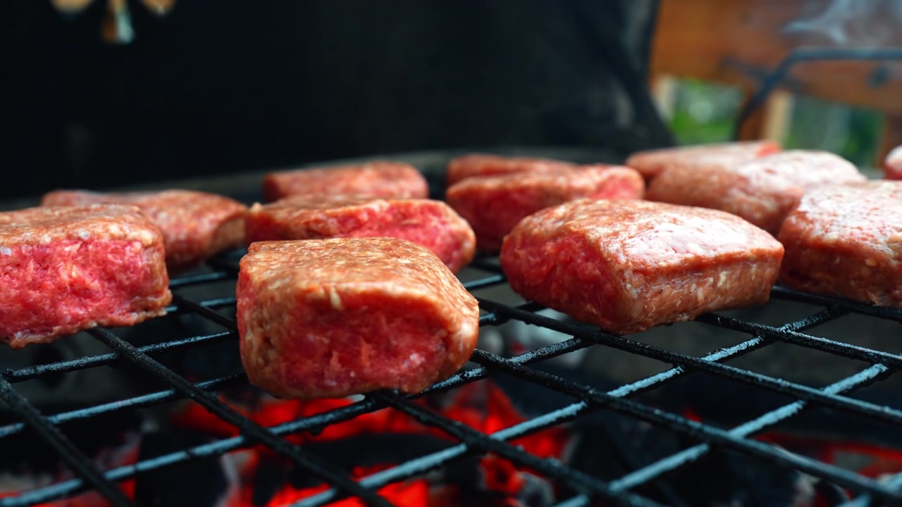 Patty daging mentah di panggangan luar ruangan dengan bara menyala. Tampilan samping sudut rendah patty daging cincang yang dipanggang di rak logam di atas arang panas yang membakar di pengaturan taman