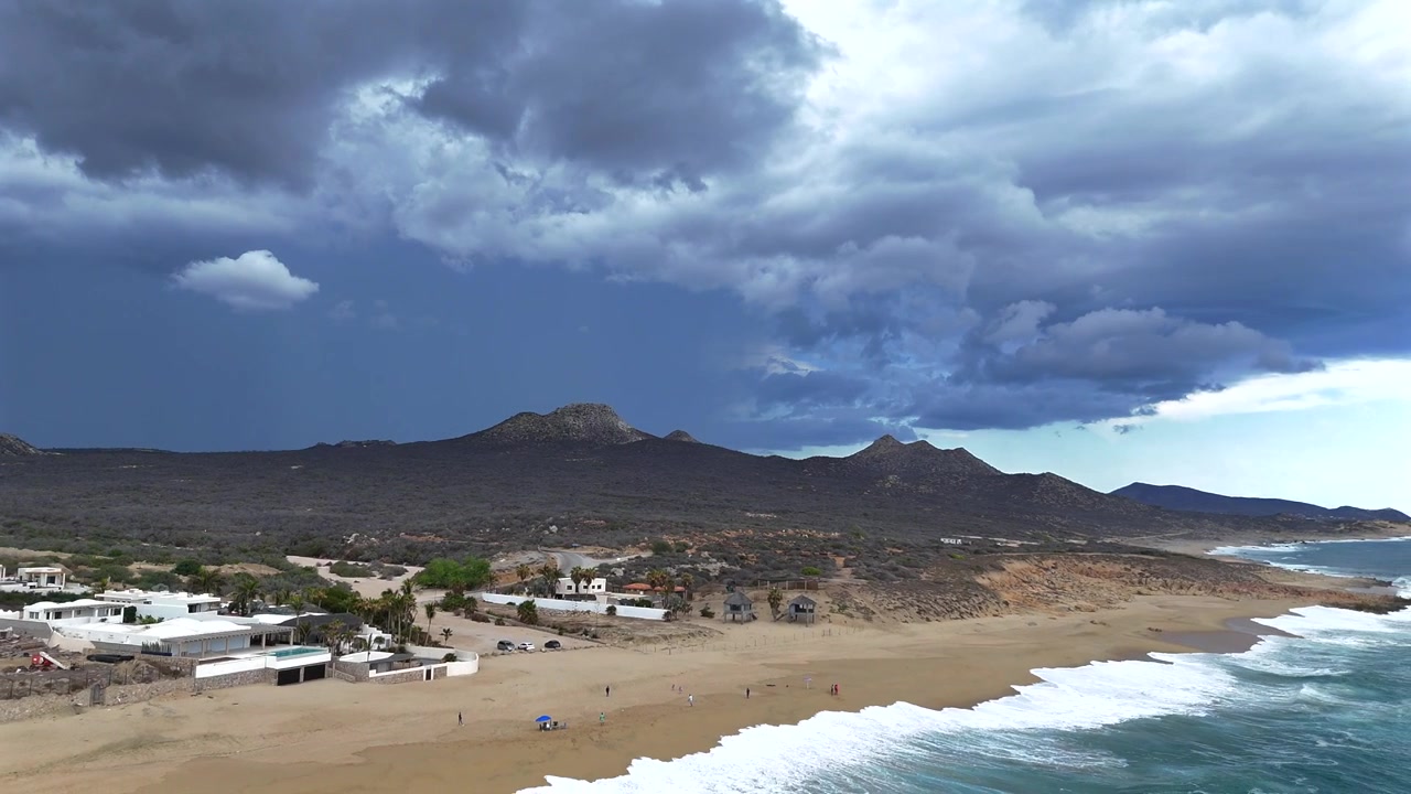 El dron se aleja mientras mira hacia tierra, retrocediendo sobre el océano para revelar la playa debajo, con nubes de tormenta oscuras acumulándose sobre las colinas orientales de La Laguna en la distancia.
