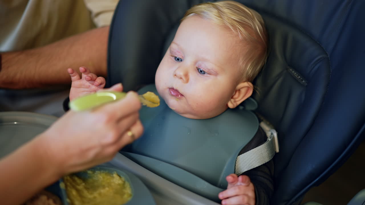 Healthy little baby boy in a blue bib is fed from spoon. Close up portrait of a child having meals.
