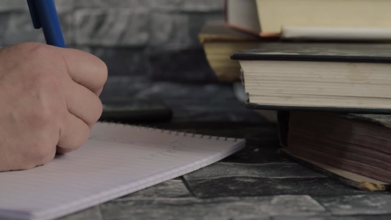 Hand writing on notepad next to pile of books close up panning shot