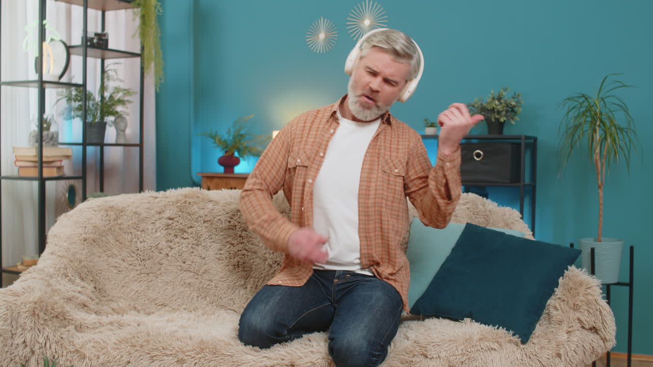 Mature man dancing rocknroll on sofa with wireless headphones playing imaginary guitar at home