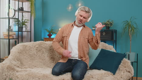 mature man dancing rocknroll on sofa with wireless headphones playing imaginary guitar at home