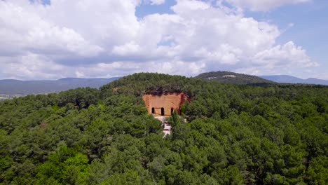 aerial-view-with-drone-of-a-hill-full-of-trees,-a-clear-and-orange-rocky-quarry-named-"Mine-de-Bruoux-",-cloudy-sky