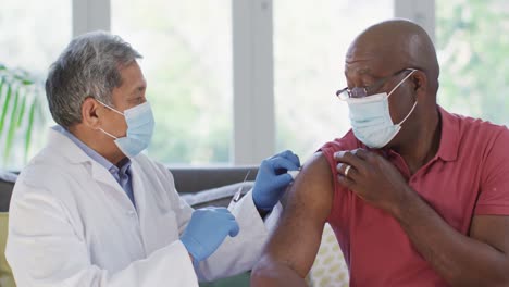 Male-doctor-wearing-face-mask-vaccinating-african-american-senior-man-at-home