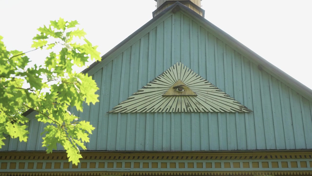 Daylit wide shot of a historic wooden building’s gable featuring an ornate All-Seeing Eye symbol. Sunlight filters through green leaves, casting dappled shadows on blue-painted wood.