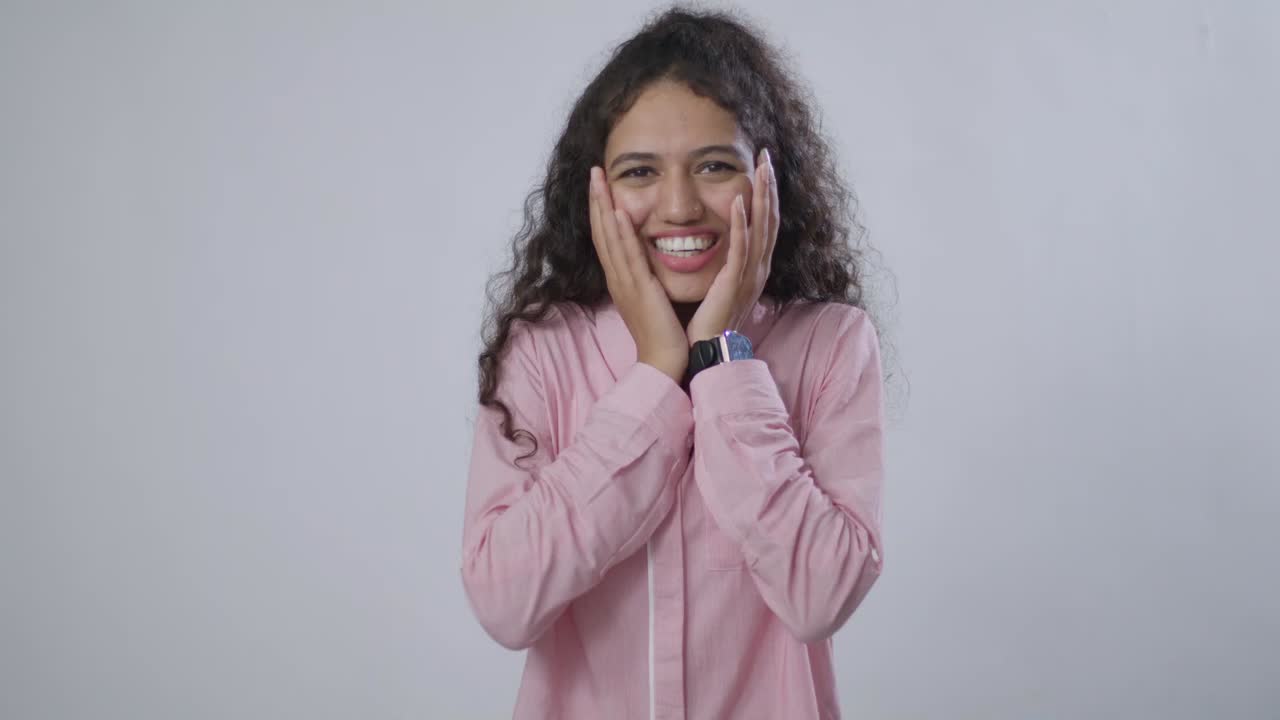 A South Asian woman with curly brown hair is smiling happily with her hands on her cheeks in surprise